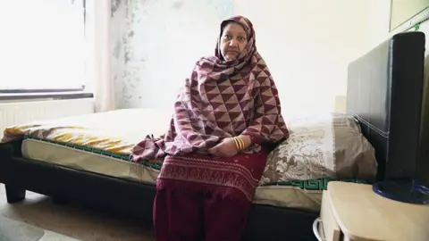 Bushra is sitting on the edge of a bed in a modest room. The bed has a dark frame with a mattress covered by patterned bedding in shades of yellow, gray, and green. Bushra is wearing a maroon outfit with a shawl featuring a red and beige geometric triangle pattern and gold bangles on the wrist. A small wooden bedside table with a blue item on top is visible to the right, and the wall behind the bed shows signs of peeling paint and discoloration.