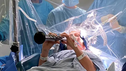 PA Media Denise Bacon lying in a hospital bed in a blue gown playing the clarinet
