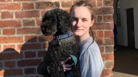 Family handout Cerys Lupton-Jones holding a dog. She is a young woman with light brown hair tied into a thin ponytail and wearing a grey shirt. She is standing in front of a brick wall and holding a medium-sized black curly haird dog