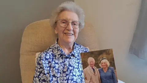 Stephanie Bell An elderly woman, 100-year-old, Jean Aitken smiles into the camera, holding her 100th birthday card from the King, which has a picture of King Charles and Queen Camilla on it. She has white hair, and is wearing a blue blouse.