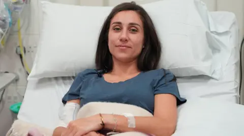 Lucy North/PA Media Emily Henders smiles at the camera while lying in a hospital bed, propped up by pillows. She has a bandage on her right arm over a cannula and a plaster on her left wrist. She wears a petrol blue t-shirt and has long brown hair.