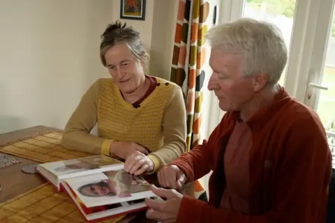 Rebecca Lupton and Dave Jones sit at a table looking at photographs of their daughter Cerys. Rebecca has grey hair with darker streaks. It is tied up and she is wearing a yellow knitted jumper. Dave has short white hair and is wearing a orange/red cardigan and a pinkish red shirt.