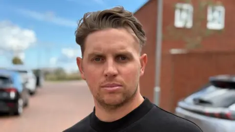 BBC A close-up on a man's face and neck. He has brown hair and stares into the camera. The background is blurred but a red-brick house is visible behind him, as are several cars on a drive.