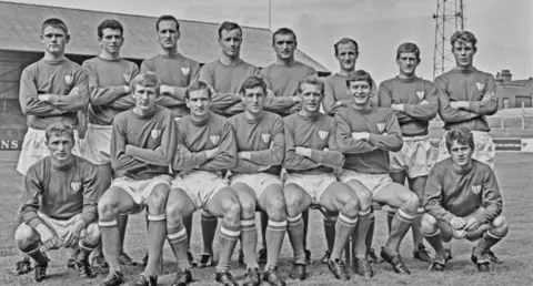 Getty Black and white group photograph of 15 football players. Half are standing up, the rest sitting down. They have their arms folded. They are on the pitch at a football ground. 