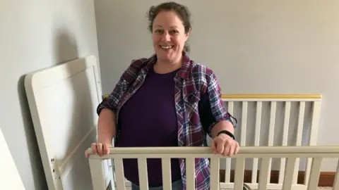 Lori Quate Jacqui Hunter, who is heavily pregnant. She is wearing a checked shirt and smiling at the camera is holding the sides of a baby cot that is being assembled