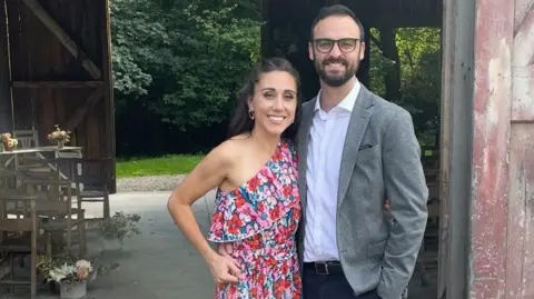 Lucy North/PA Media EMily and her husband smile into the camera. She is wearing a pretty pink floral dress that is off the shoulder and her husband wears a grey blazer. He has a beard and glasses