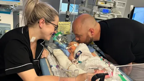 Family handout Tom and Becky Williams are perched over Mabel who is surrounded by wires & machines in a hospital. Mabel is lying down on a bed. Tom is kissing her on the head while Becky smiles.