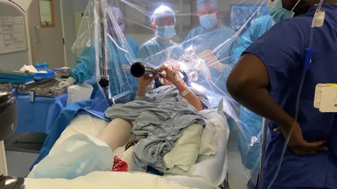 PA Media Denise Bacon lying in a hospital bed in a blue gown playing the clarinet