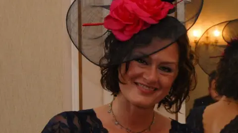 Family photo Maria Morris, who died at Bethlem Royal Hospital in 2021, is standing in front of a mirror at a wedding smiling. She is wearing a black dress, and a black and pink fascinator. 