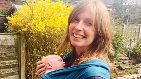 Helen Ledwick Helen smiling at the camera with a baby in a sling