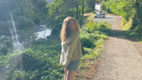 Emily wears denim shorts and a jumper, and is standing on a path beside a stream, bordered by greenery, while the sun shines in the background