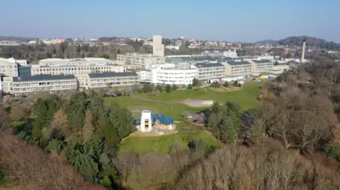 An ariel view of a white and grey building surrounded by woodlands with the city of Dundee in the background. 
