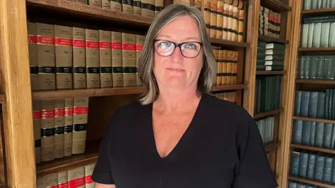 Jacquie Russell has shoulder-length grey hair and dark rimmed glasses. She is wearing a black dress and looking at the camera. Behind her are shelves filled with books.