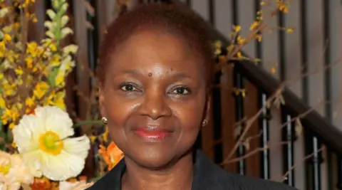 A woman with dark hair in front of a striped background and some flowers. She is smiling at the camera.
