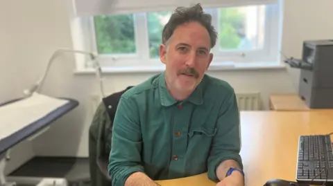 A man in a green shirt sits at a desk in a GP surgery. A keyboard can be seen to his right
