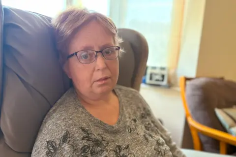 A woman with short blonde hair and black glasses sits in a brown leather chair and looks at the camera. She wears a thin grey jumper with flowers on it.