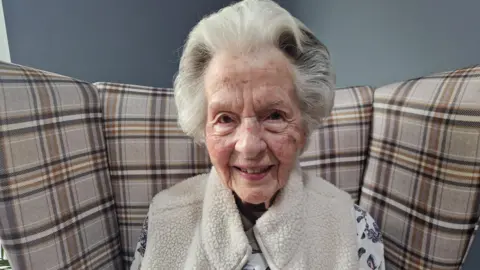 Murrayfield Care Home An elderly woman, 104-year-old, Mary Smith smiles into the camera. She has white hair, with dark streaks and is wearing a cosy cream gilet over a patterned top.