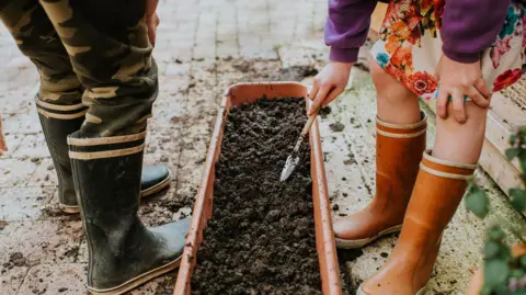 Getty Images Focus is on the wellies of two children - a girl and a boy, using a small trowel to plant seeds in a long planter trough.