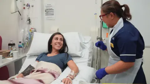 Lucy North/PA Media Emily lies in the hospital bed looking up at a female nurse who stands next to her. They are laughing together with wide smiles. The nurse wears blue rubber gloves and a white plastic apron, and has her hair tied back in a ponytail. Lucy has tubes going into her arm and hand, and there is a drip stand next to her bed.