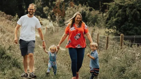 Family handout A man and woman holding the hands of their two children, a young girl and boy, walking along a grass path. The man is wearing a white top and grey shorts with trainers and glasses and smiling at the camera. A little girl is wearing blue dungarees. A woman is looking down at her little boy and has long blonde hair and an orange flowery top and blue jeans. The little boy is standing to the side and smiling at the camera.