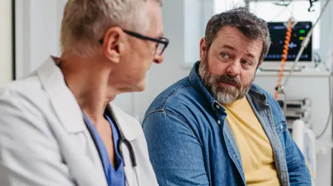 Getty Images A doctor wearing scrubs and a white coat talks to a middle-aged man wearing a yellow t-shirt and denim shirt. 