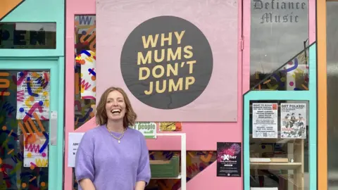 Helen Ledwick Helen wearing a purple jumper and smiling in front of a poster which says 'why mums don't jump'