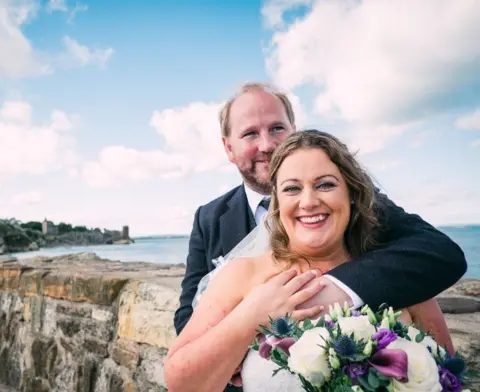 Fern Photography Lori and Jacqui. He is in a light grey formal suit and has his arms around Jacqui who is in a wedding dress. She is smiling and holding flowers. They are pictured next to a stone harbour wall next to the North Sea. 