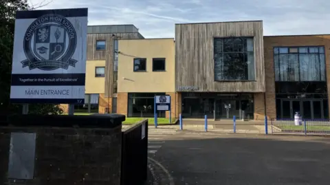 BBC The front of a school building with a mix of facades - some wooden slatted with large windows and some stone and brick - and a driveway leading up to it. There are blue bollards directly in front of a brown reception area. A sign at the entrance reads Congleton High School Main Entrance.