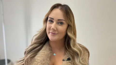 Lizzie has long, wavy, highlighted brown hair. She is wearing a faux fur gilet and black top, with a necklace and is smiling at the camera. She is sat in a white clinical treatment room. It is a head and shoulders shot of her. 