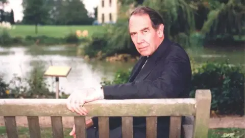 Royal Papworth Sir Terence English sitting on a bench, looking back towards the camera. His left hand is resting on the back of the wooden bench. He is wearing a dark jacket. There is a lake and a house in the background.
