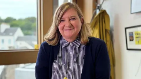 Jenni Minto wearing a dark cardigan over a striped button-up shirt, standing indoors near a window with a view of buildings and greenery outside; a yellow coat is hanging on the wall in the background, and framed artwork is partially visible.