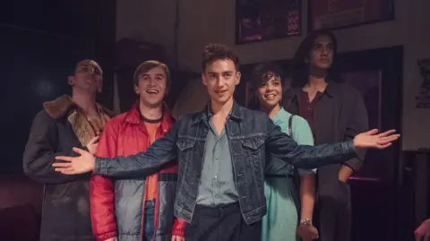 Channel 4 This photograph includes five friends. From left to right we have a man in a black jacket with a fur collar, a man in a red puffer coat, a man in a denim jacket, a woman in a blue dress and finally (on the far right) a man in a grey jacket. They're all looking in different directions. 