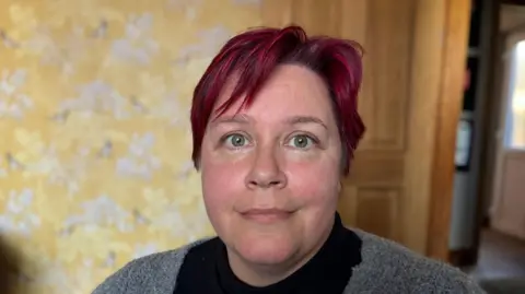 Lady with red hair and greenish-grey eyes, wearing a black top and grey cardigan, looks straight ahead. Behind her in the background is some flowery, yellow wallpaper which is blurred and a wooden door.
