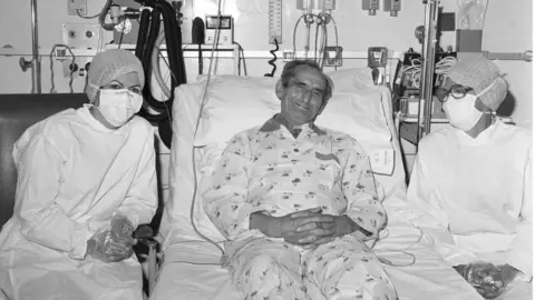 PA Media A black and white photo of Keith propped up in a hospital bed, looking at the camera and smiling. He wears a hospital gown or pyjamas, and has various tubes attached. On his left and right sit two women wearing white hospital gowns and plastic gloves. There are various hospital instruments and tubes behind him.