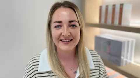 Hannah has long, straight blonde hair and is smiling at the camera. She is wearing a cream and black striped top, with a white collar. She is stood next to shelves displaying products in a clinic. It is a head and shoulders shot.
