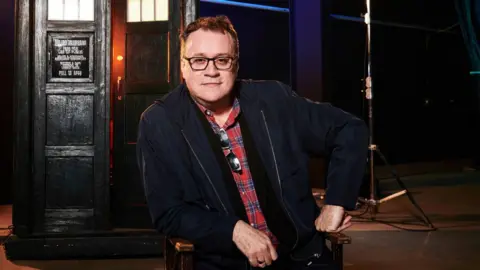 Ray Burmiston In this photograph Russell T Davies is wearing a red and blue checked shirt with a black jacket. It's a professional photograph taken on the set of Dr Who. You can see the Tardis in the background. He has dark hair and wears black glasses. 