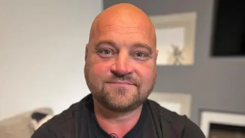 Offshore worker Phil Perry, who has a bald head and is wearing a black T-shirt, is smiling at the  camera. He has tattoos on arms.