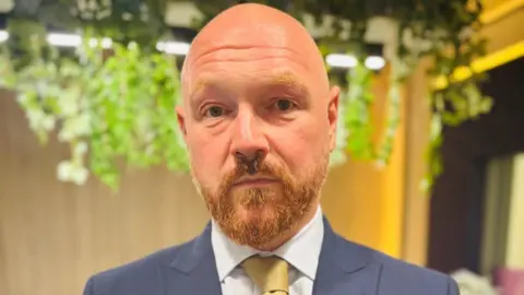 Graham Skinner, a man with a beard, wearing a blue suit, white shirt and yellow/gold tie looking at camera.