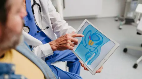 Getty Images A doctor wearing a white coat with a stethoscope around his neck is pointing at a picture of a man's prostate on a tablet, while talking to a male patient with a greyish bear who is blurred in the foreground.
