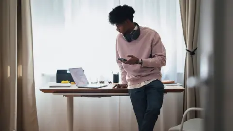 Getty Images Stock photo shows a young man looking at his phone while standing next to his laptop which is on a desk next to him inside next to a window.
