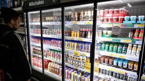 Getty Images A man with dark hair stands in front of a large supermarket fridge containing shelves of milk-based drinks, such as flavoured milkshakes and milky coffees like lattes, cappucinos and iced coffees.