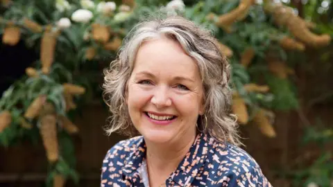 Supplied A smiling Alex Allan looks directly at the camera as she is photographed outside before a plant. She has grey hair and is wearing hoop earrings and a multi-coloured shirt. 