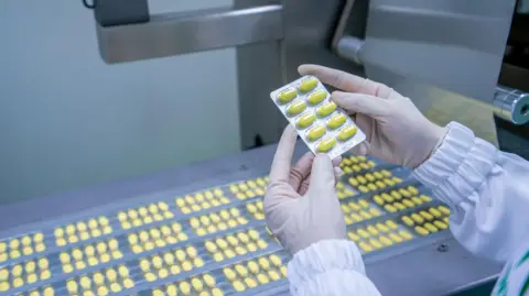 Getty Images Gloved hands hold a tray of yellow tablets, above rows of yellow tablets on a conveyor belt.
