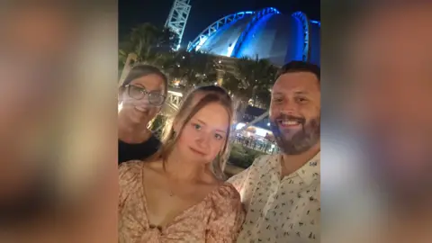 Family handout A young woman is standing in the middle of her mum and dad smiling at the camera. She has a pink dress on and mousey blonde hair. Her dad is on the right and has a white shirt with dark blue small patterns. Her mum is on the left with a black top. It is a head and shoulders selfie shot of them.